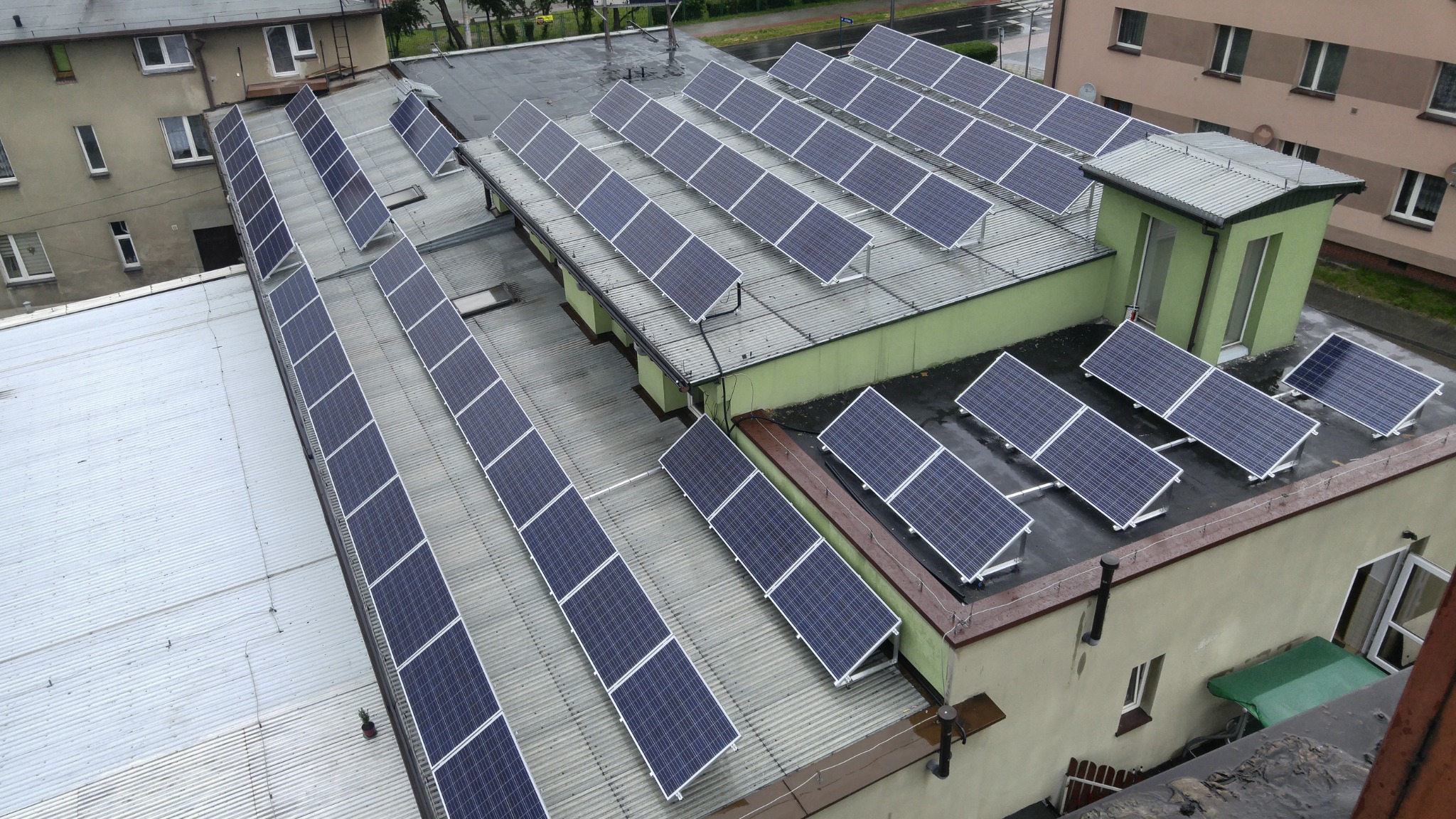 Widok z góry na instalację paneli słonecznych na dachu budynku z różnymi rodzajami pokrycia, w pochmurny dzień.