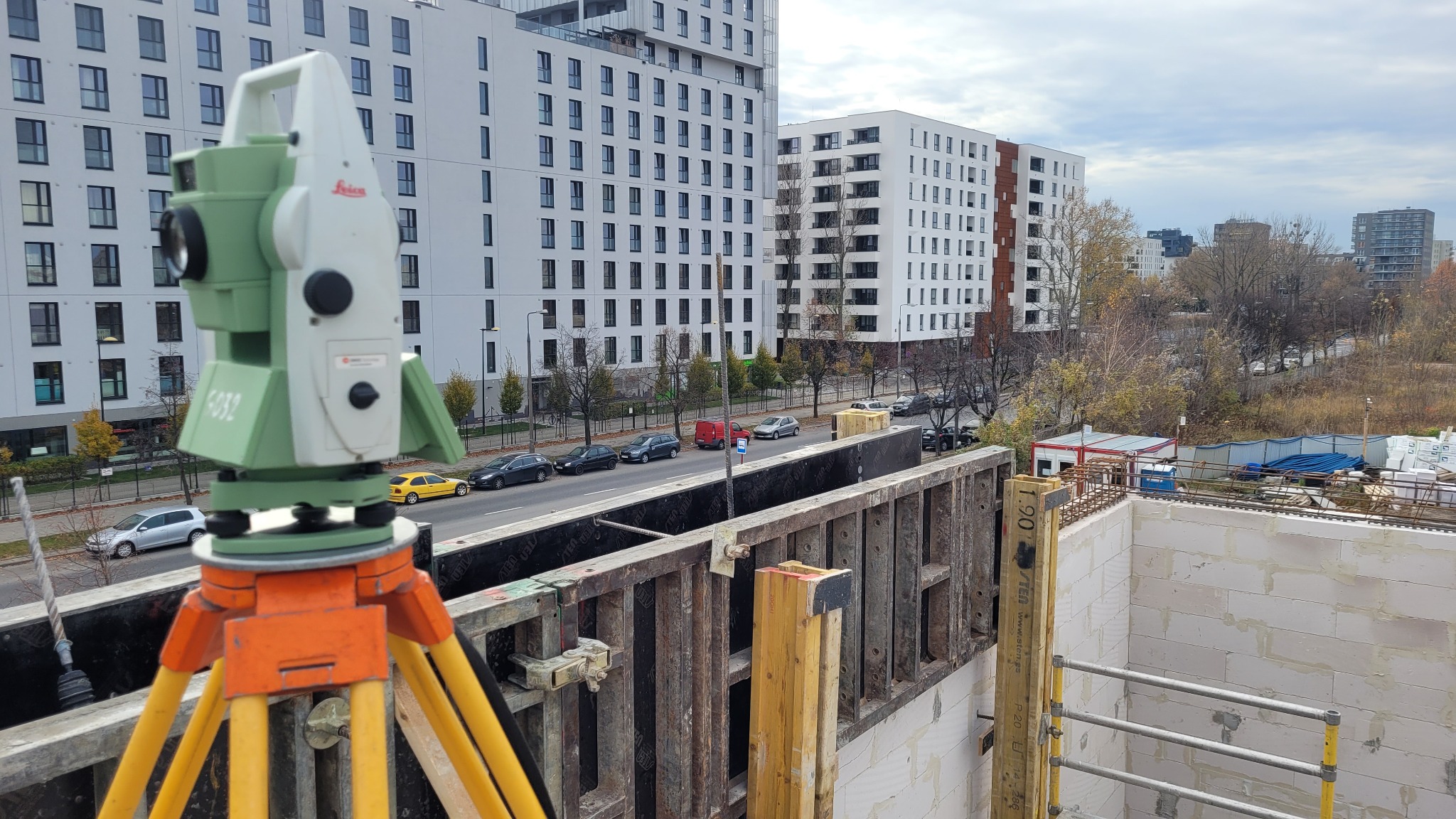 Zielono-biały teodolit Leica na pomarańczowym statywie, ustawiony na budowie bloku mieszkalnego, z widokiem na ulicę i zaparkowane samochody.