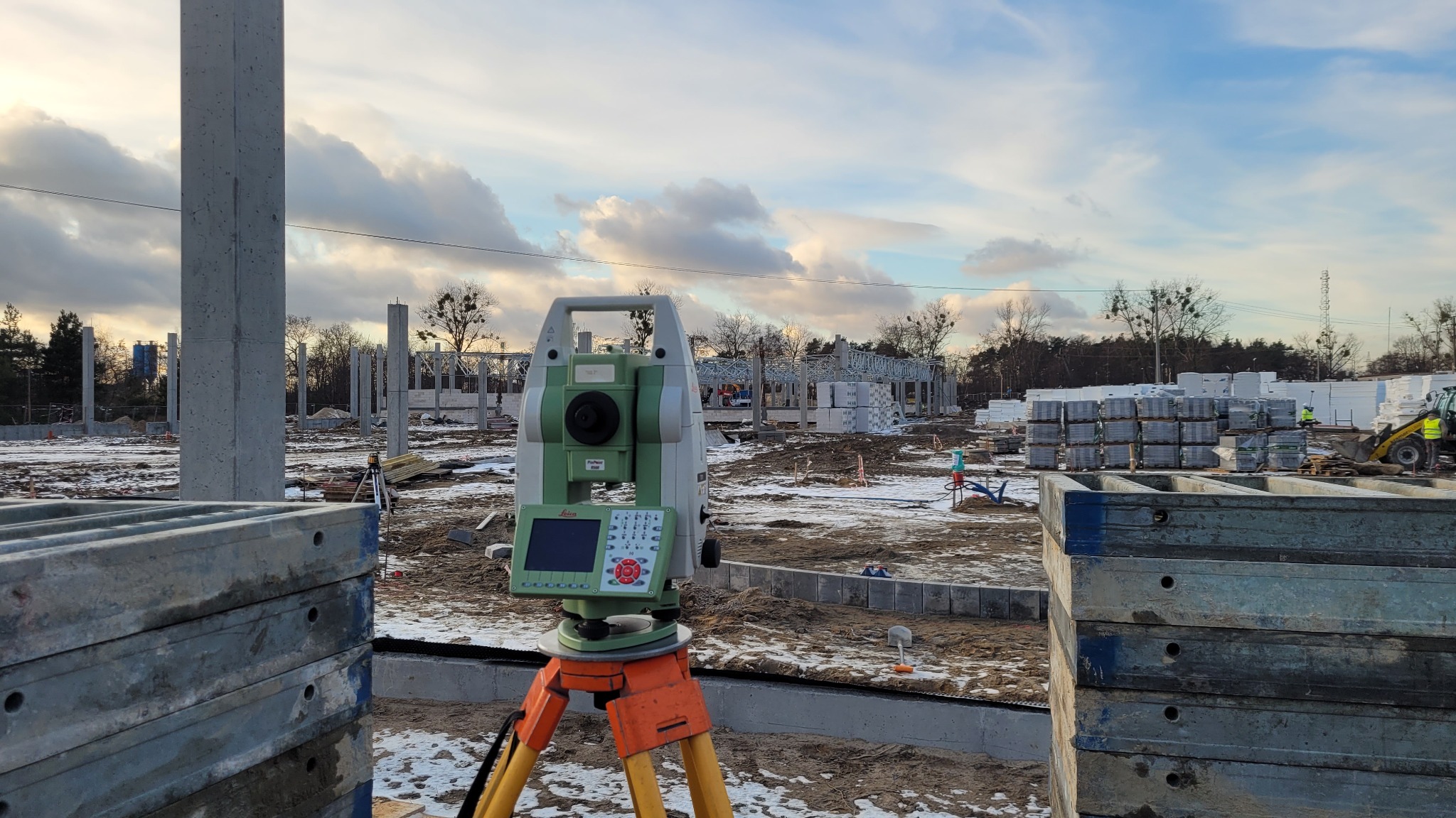 Zielony teodolit marki Leica na pomarańczowym statywie, ustawiony na placu budowy z widocznymi elementami konstrukcyjnymi i stertami materiałów budowlanych w tle, przy pochmurnym niebie.