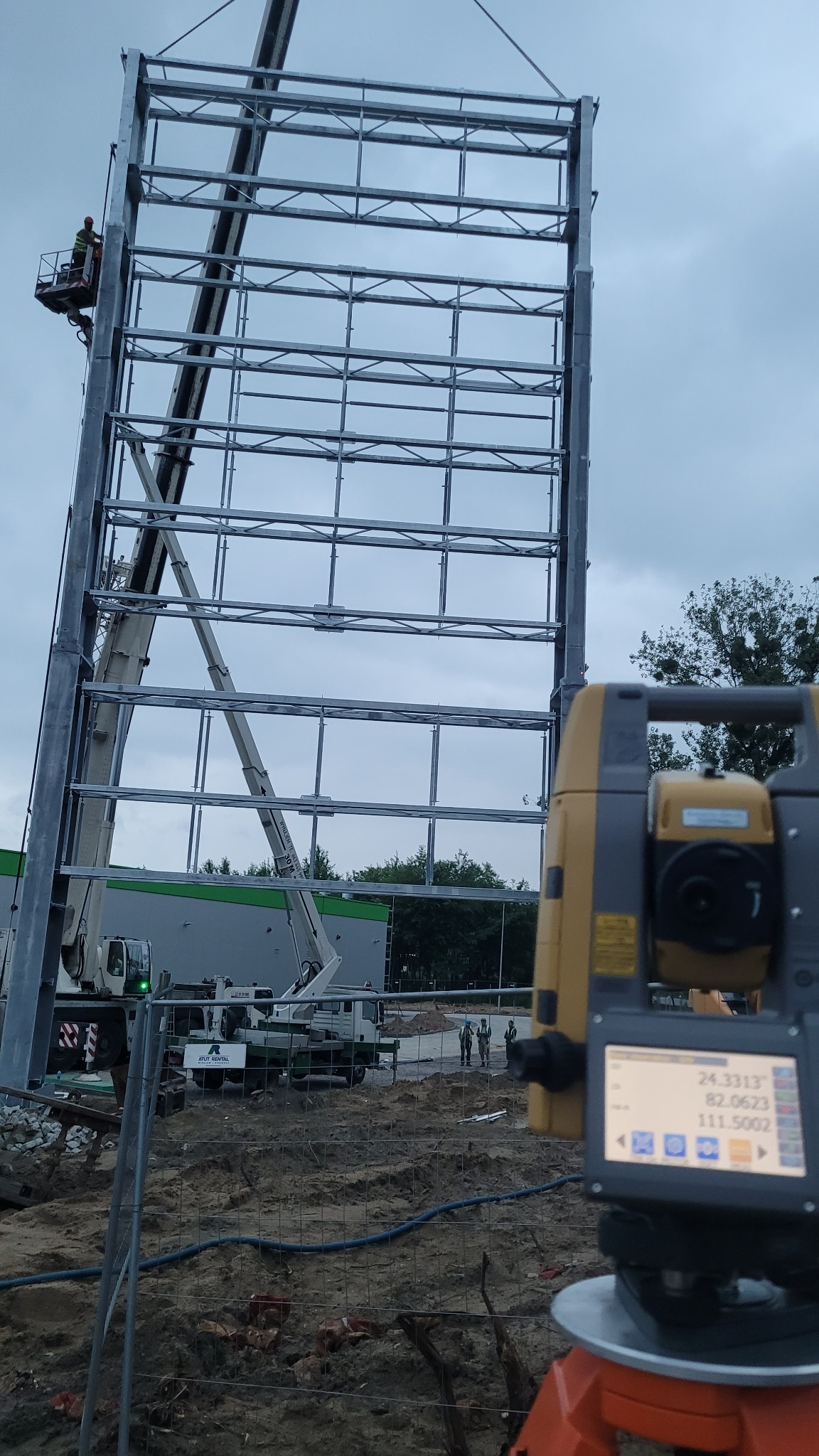 Montaż stalowej konstrukcji hali przemysłowej kontrolowany teodolitem, widoczny pracownik w koszu podnośnika i dźwig