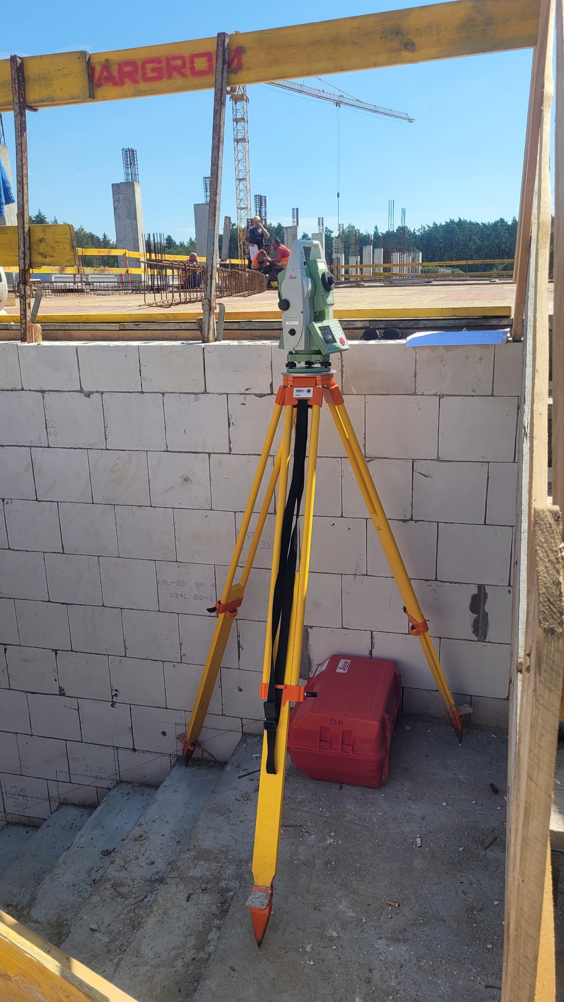 Tachimetr Leica na żółtym statywie ustawiony na placu budowy w Warszawie, widoczne białe bloczki, czerwona skrzynka i w tle konstrukcja budynku oraz żuraw budowlany.