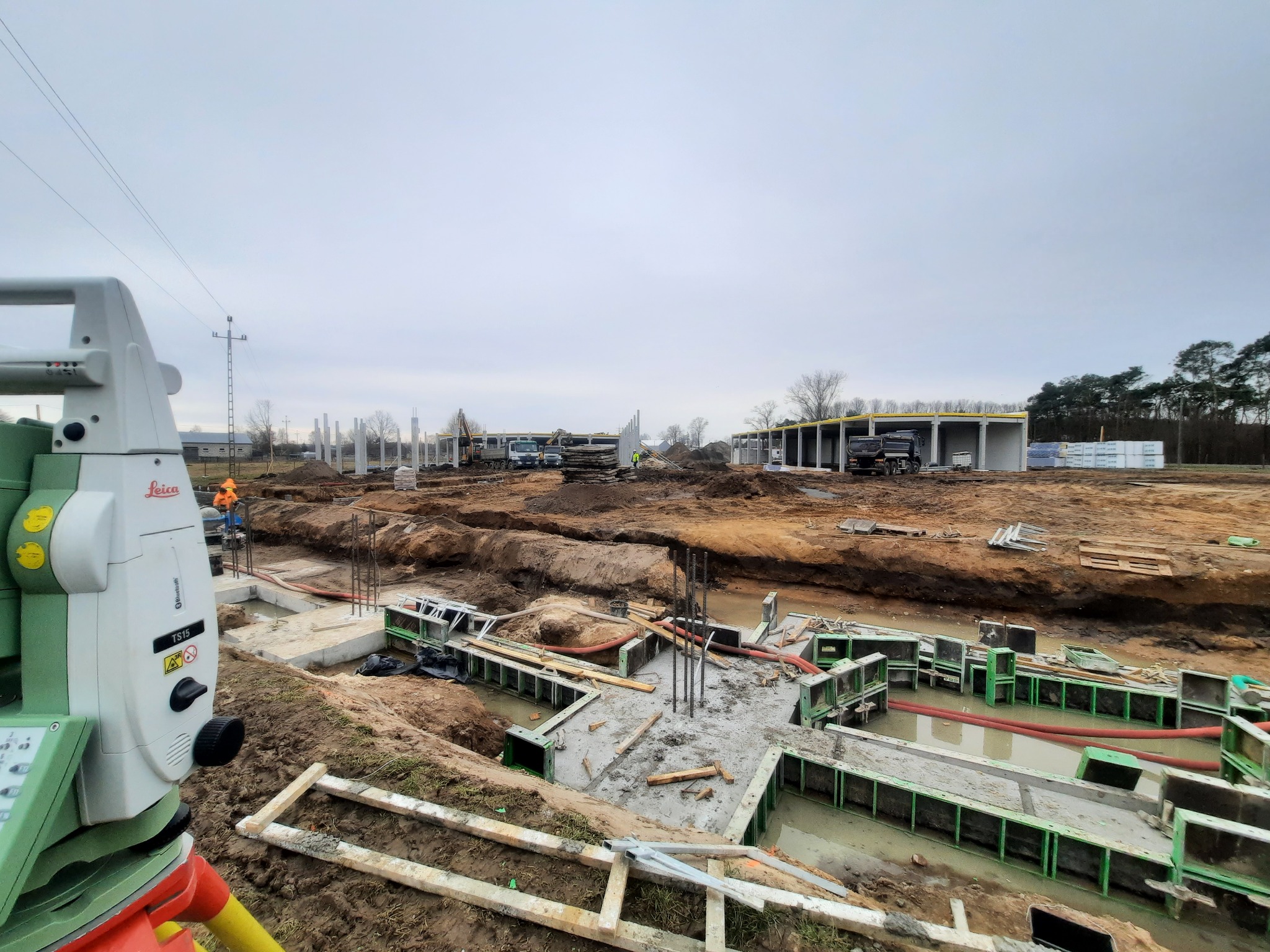 Pomiar Leica TS15 na budowie fundamentów hali, widoczne zbrojenia i szalunki, w tle betonowe słupy konstrukcyjne i ciężki sprzęt.