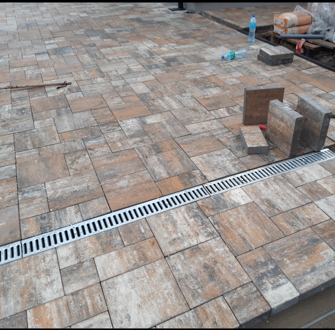 Ułożona kostka brukowa w odcieniach brązu i szarości, z widocznym odpływem liniowym. Część kostki jeszcze nieułożona obok, widoczne butelki z wodą i worki z materiałem.