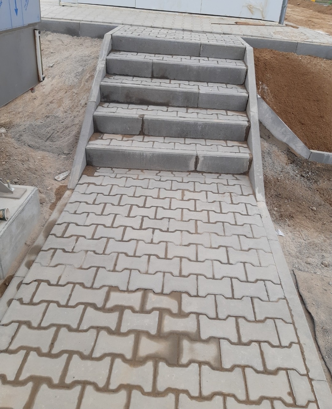 Ułożona szara kostka brukowa prowadząca do schodów, widoczne ślady wilgoci na powierzchni, obok piasek i elementy konstrukcyjne budynku.