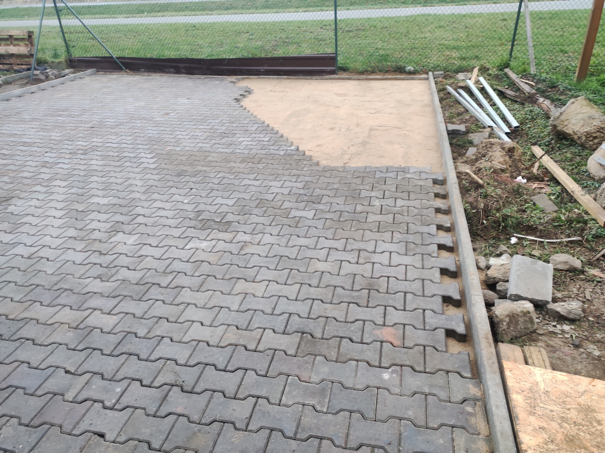 Układanie szarej kostki brukowej typu 'I' na podsypce piaskowej, widoczne niedokończone krawędzie i przygotowane materiały budowlane.
