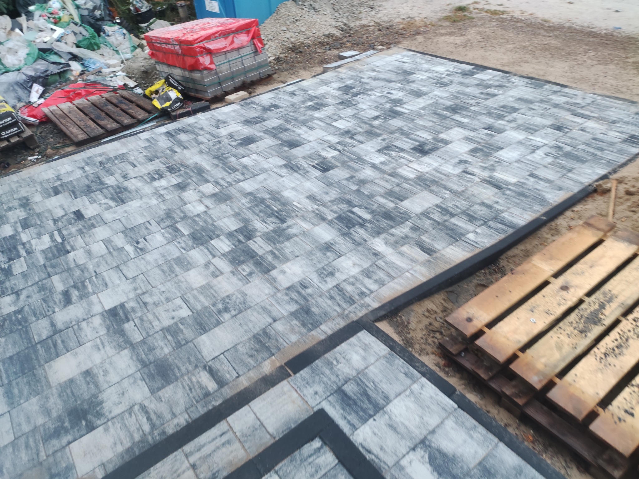 Ukończona powierzchnia z ułożonej szarej kostki brukowej o nieregularnym wzorze, obramowana ciemnoszarą kostką, z paletami kostki czekającymi na wykorzystanie w tle.