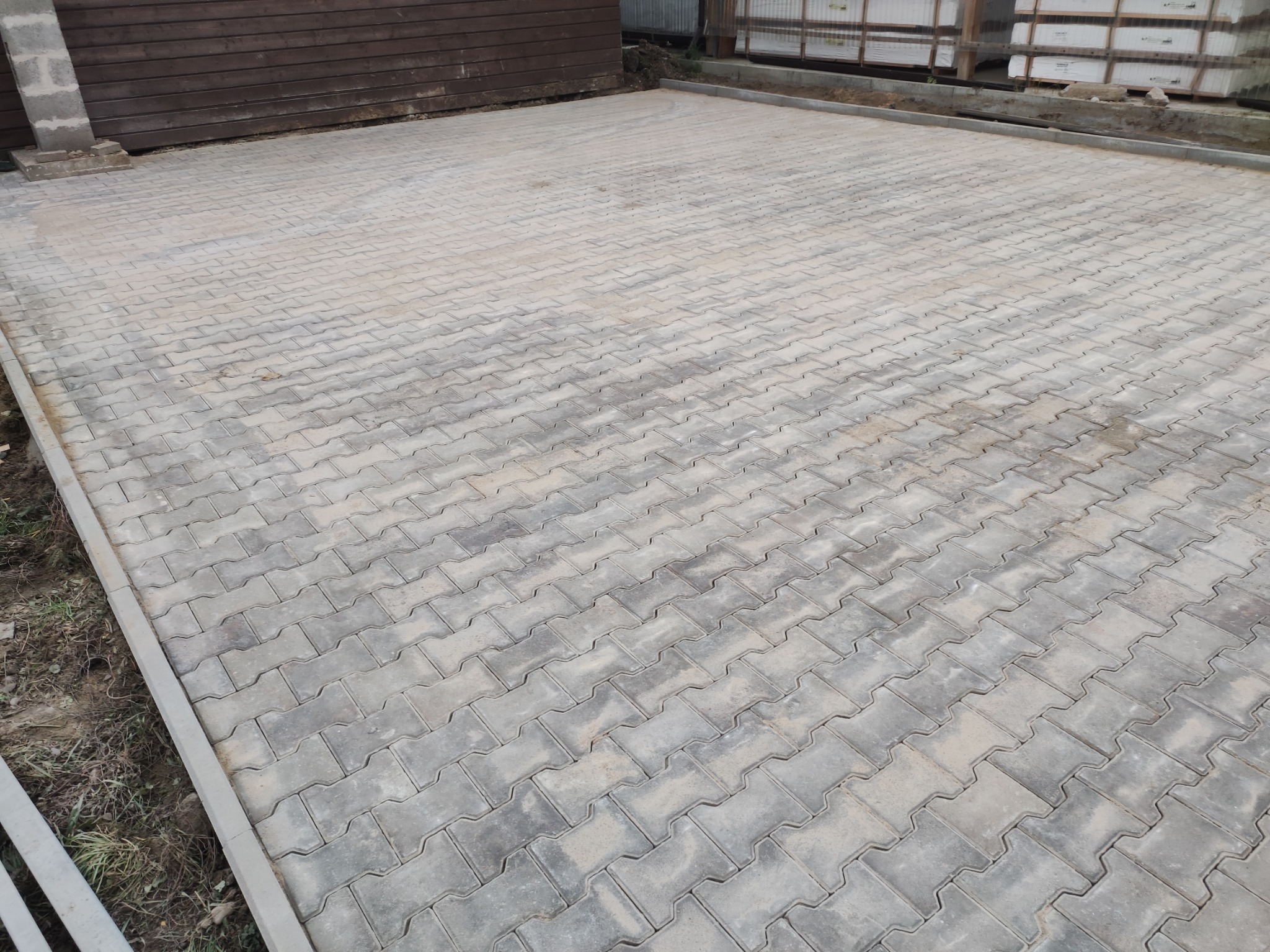 Ułożona szara kostka brukowa typu 'kość', ograniczona szarym obrzeżem, widoczny fragment drewnianego budynku i palety z kostką w tle.
