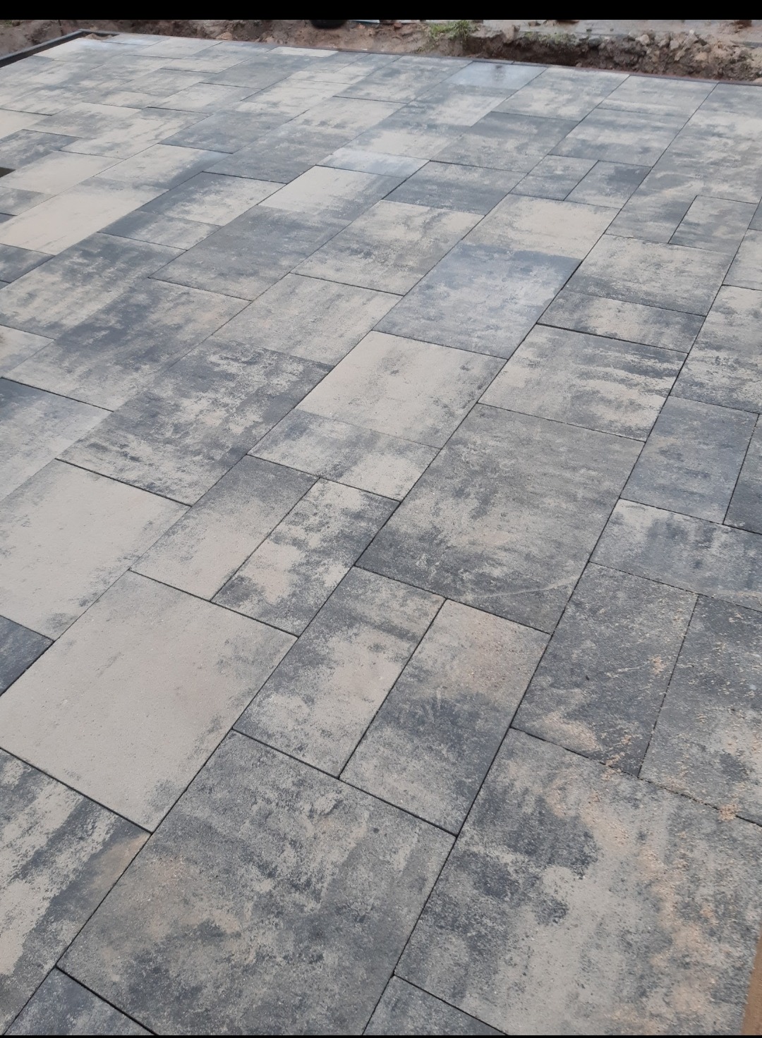 Ułożona nawierzchnia z kostki w odcieniach szarości i beżu, wzór prostokątny i kwadratowy, widoczne nierówności kolorystyczne na poszczególnych elementach, świeżo ułożona z widocznymi śladami piasku.