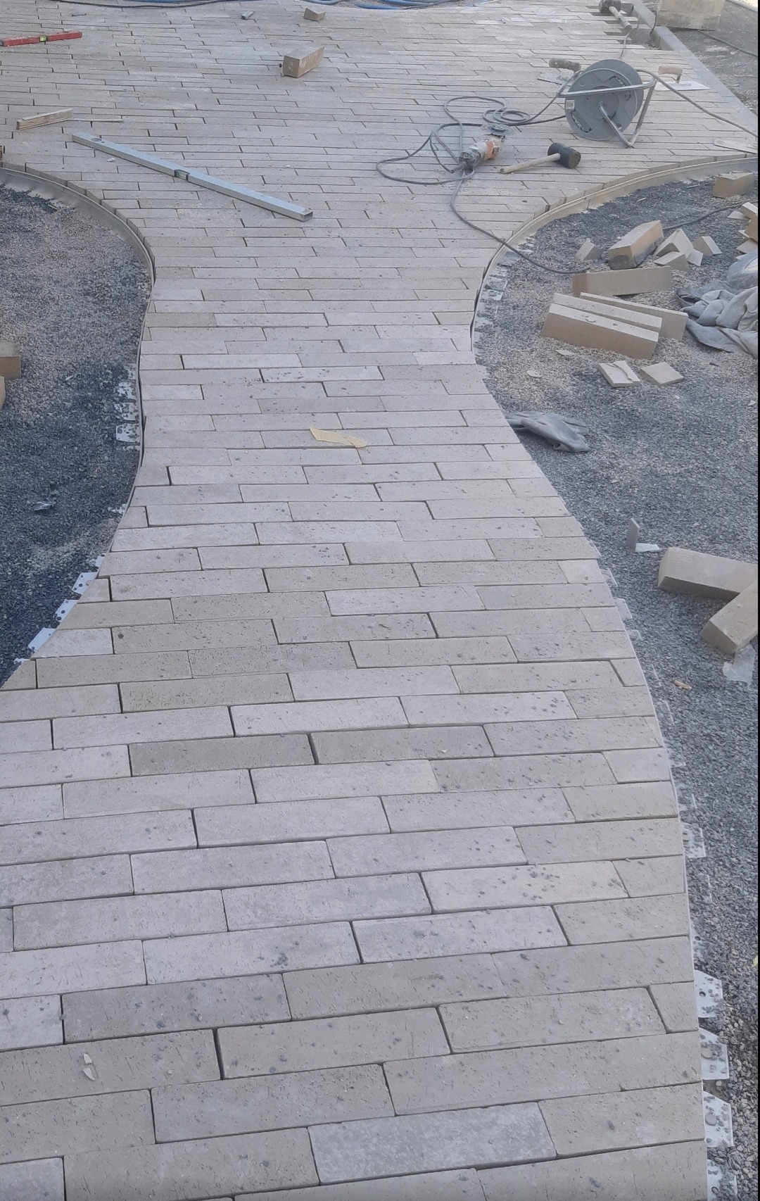 Wężykowaty chodnik z jasnych, prostokątnych kostek w trakcie układania, widoczne narzędzia: poziomica, szlifierka kątowa, młotek, oraz zapas kostek i podsypka.