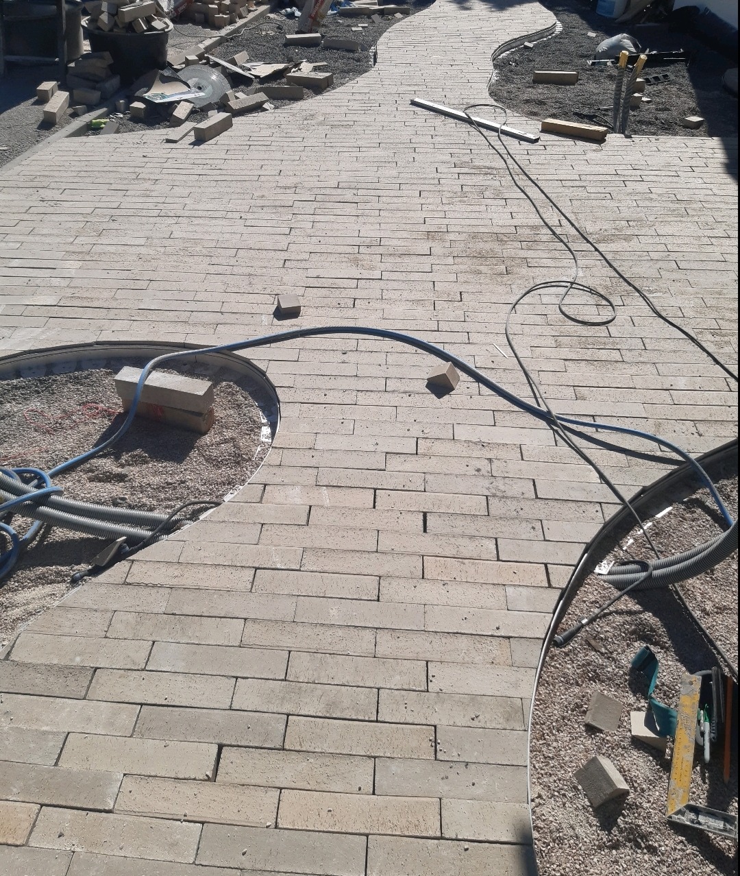 Ułożona częściowo ścieżka z prostokątnej, beżowej kostki, widoczne kable, piasek i narzędzia w trakcie prac wykończeniowych.