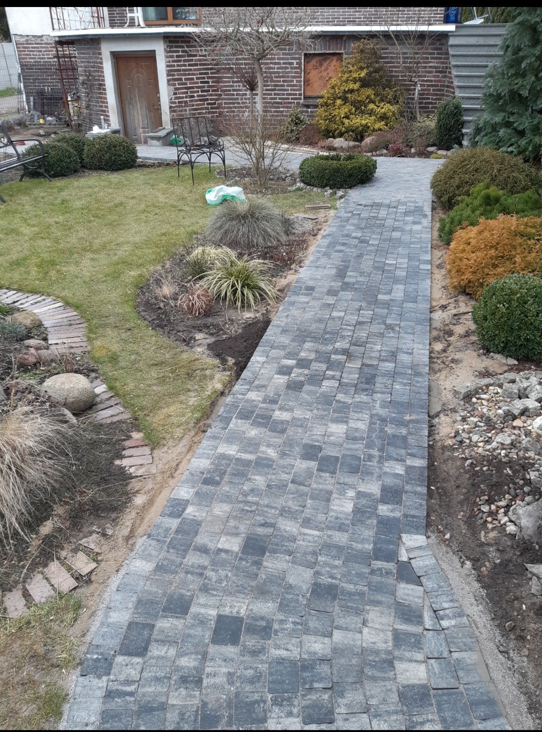 Ścieżka z szarej kostki brukowej prowadząca przez ogród z krzewami i trawami ozdobnymi, widok na dom z cegły w tle.