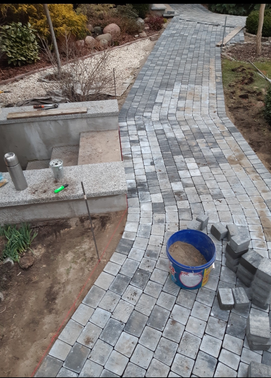 Układanie ścieżki z kostki brukowej w odcieniach szarości, widoczny fragment ogrodu, narzędzia i wiadro z piaskiem w trakcie realizacji.
