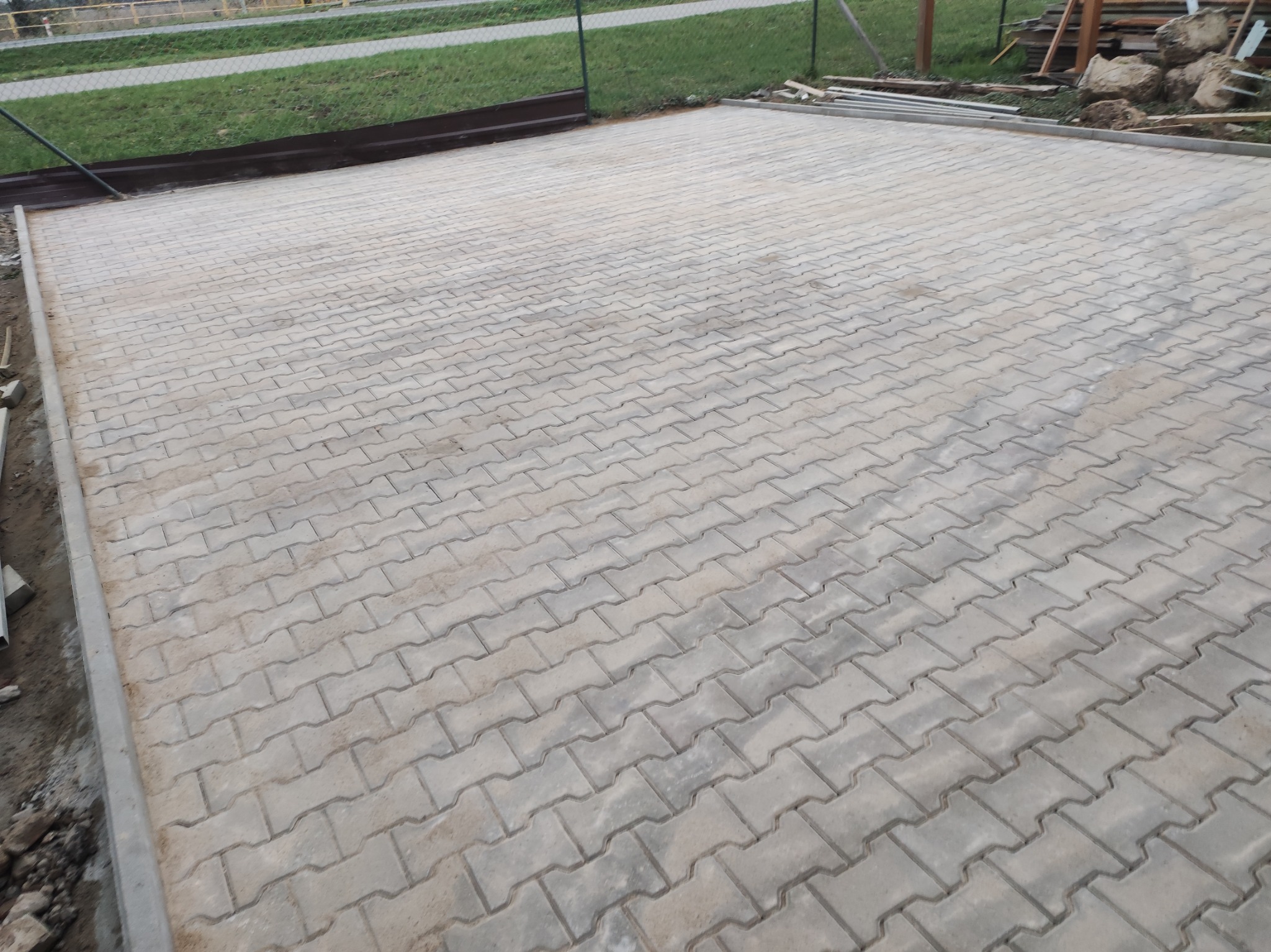 Ułożona szara kostka brukowa na posesji, widoczne obrzeża, trawnik i ogrodzenie w tle.