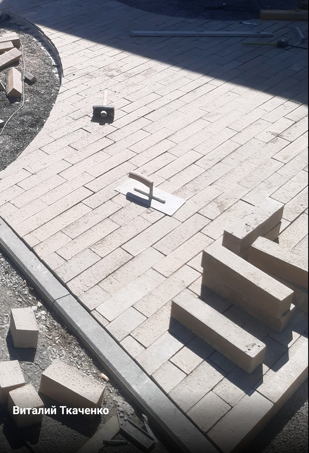 Układanie jasnej kostki w jodełkę, widoczna kielnia, gumowy młotek i nieułożone kostki na obrzeżach ścieżki.