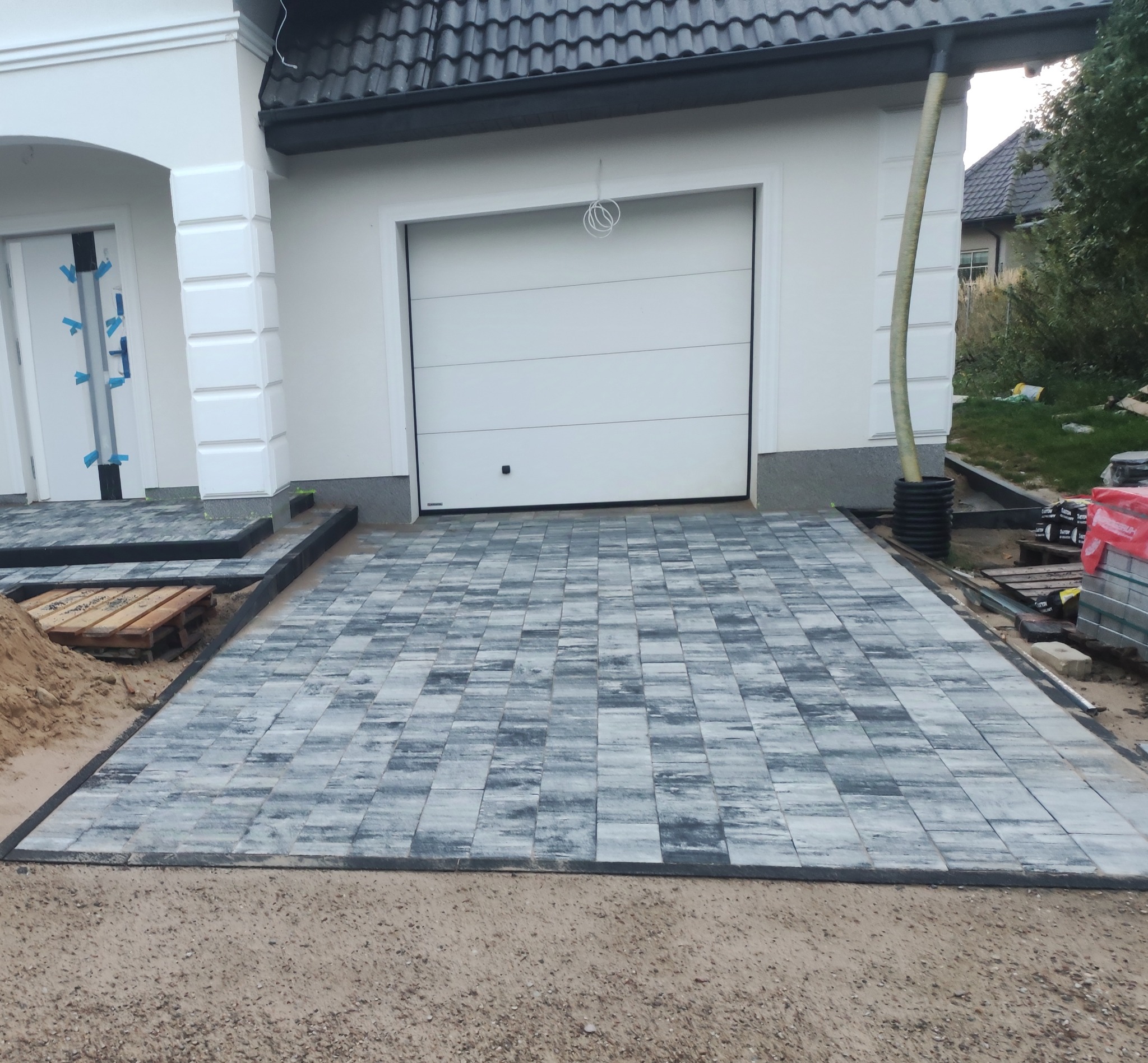 Ułożona szara kostka brukowa przed nowoczesnym domem z białą elewacją i garażem, widoczne palety i pozostałości piasku po pracach.
