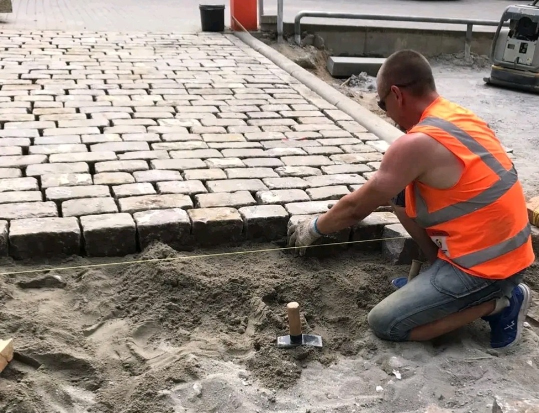 Pracownik w kamizelce odblaskowej układa kostkę granitową na podsypce piaskowej, widoczne narzędzia i częściowo ułożona nawierzchnia.