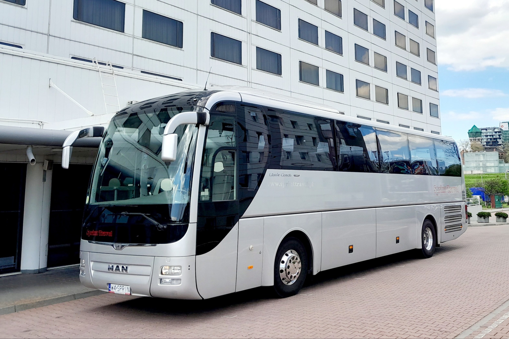 Srebrny autokar MAN z napisem Sprint Travel parkuje przed nowoczesnym budynkiem z licznymi oknami. Widoczna tablica rejestracyjna i logo firmy.