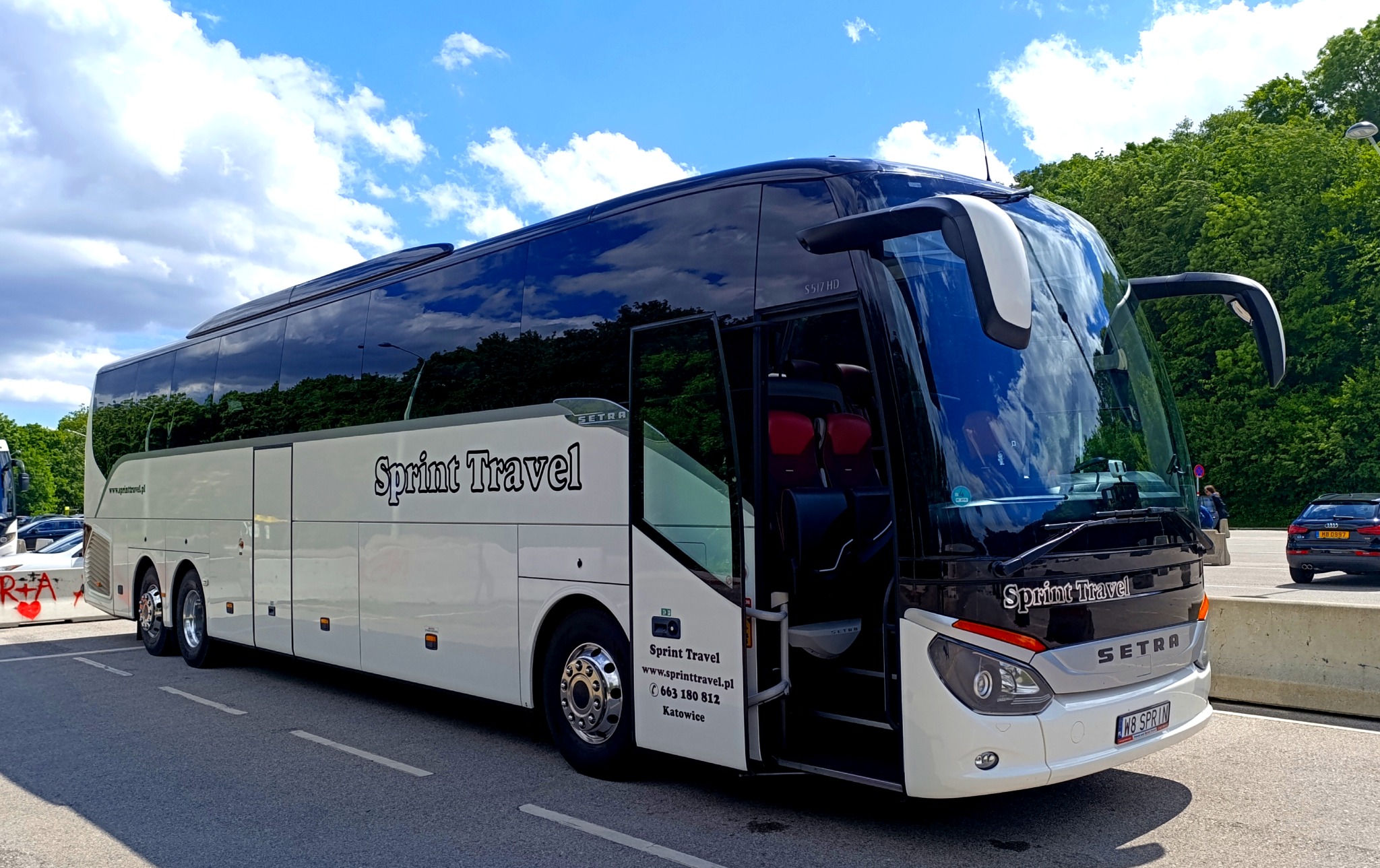 Nowoczesny autokar turystyczny Sprint Travel z otwartymi drzwiami na tle zieleni i błękitnego nieba z chmurami. Widoczny numer telefonu i adres strony internetowej.