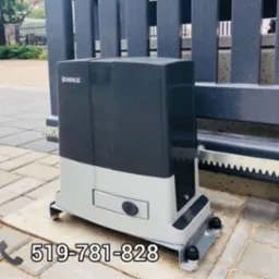 Szaro-czarny napęd do bram marki Beninca zamontowany na kostce brukowej, widoczna zębatka i fragment bramy.