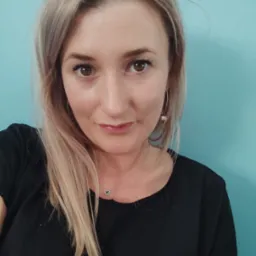 Portret kobiety z blond włosami, srebrną biżuterią i czarną koszulką na tle turkusowej ściany.