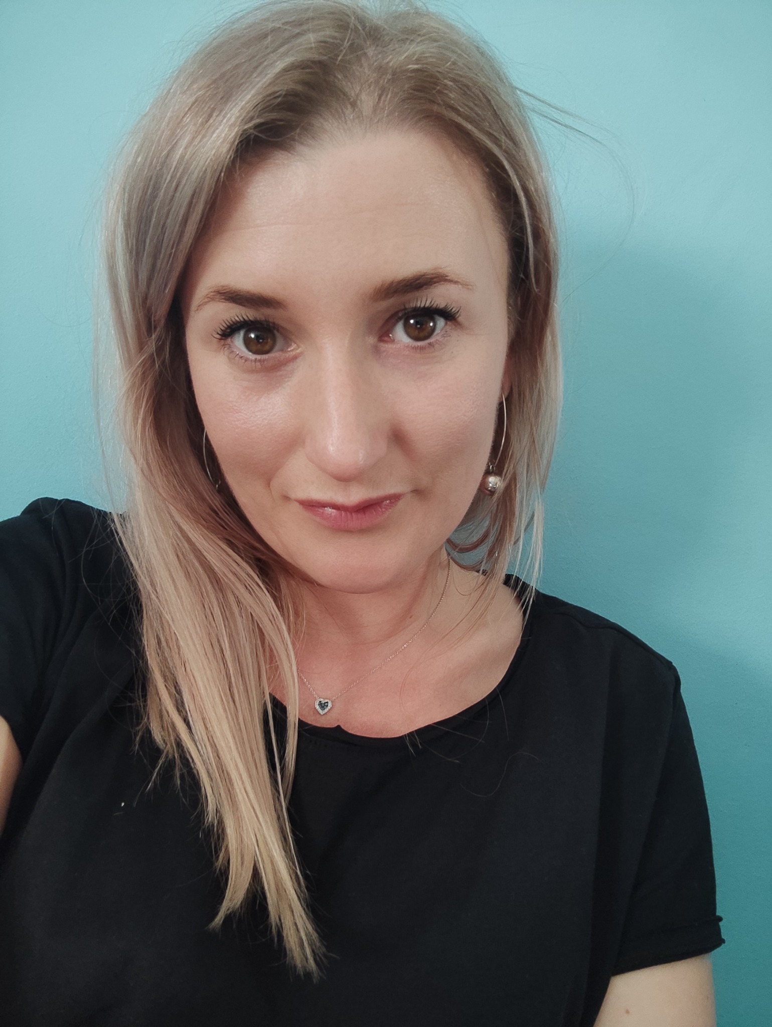 Portret kobiety z blond włosami, srebrną biżuterią i czarną koszulką na tle turkusowej ściany.