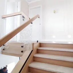 Jasne drewniane schody z dekoracyjną balustradą z ornamentowanego szkła, oświetlenie punktowe w suficie, białe ściany z subtelną sztukaterią.