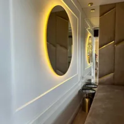 Owalne lustro z podświetleniem LED na białej ścianie z dekoracyjnymi listwami, odbijające fragment tapicerowanej ściany z geometrycznym wzorem i złotymi akcentami.