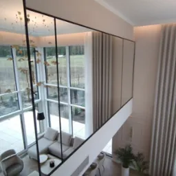 Wysokie okna z czarnymi szprosami i jasnymi zasłonami w przestronnym salonie z antresolą, widok z góry.