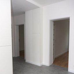 GRUPA FACHOWO SPÓŁKA Z OGRANICZONĄ ODPOWIEDZIALNOŚCIĄ - Jasne wnętrze mieszkania z szarymi płytkami podłogowymi, białymi ścianami i zabudową meblową, widok na otwarte drzwi do sąsiednich pomieszczeń z drewnianą podłogą.