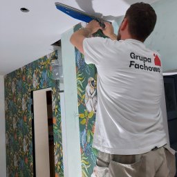 GRUPA FACHOWO SPÓŁKA Z OGRANICZONĄ ODPOWIEDZIALNOŚCIĄ - Pracownik w koszulce firmowej wygładza gładź na suficie w pomieszczeniu z tapetą w motywy dżungli.