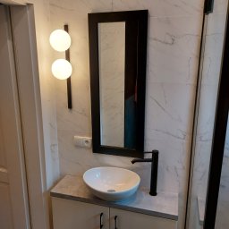 GRUPA FACHOWO SPÓŁKA Z OGRANICZONĄ ODPOWIEDZIALNOŚCIĄ - Nowoczesna łazienka z umywalką nablatową, czarną armaturą i podświetlanym lustrem na tle marmurowej ściany.