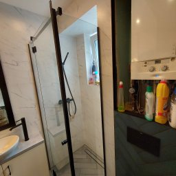 GRUPA FACHOWO SPÓŁKA Z OGRANICZONĄ ODPOWIEDZIALNOŚCIĄ - Nowoczesna łazienka z kabiną prysznicową typu walk-in z czarnymi profilami, umywalką nablatową na szafce w kremowym kolorze, marmurowymi płytkami na ścianach i podłodze ułożonej w jodełkę; obok...