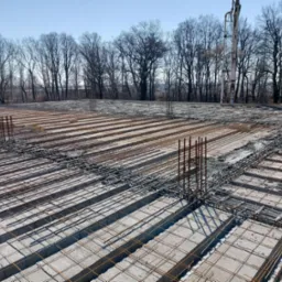 Zbrojenie fundamentów płyty fundamentowej z betonem w trakcie wylewania, widoczna pompa do betonu i drzewa w tle.