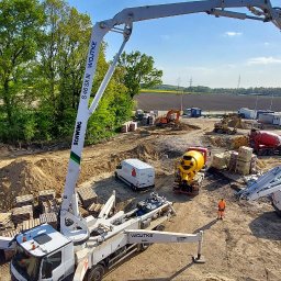 GRUPA FACHOWO SPÓŁKA Z OGRANICZONĄ ODPOWIEDZIALNOŚCIĄ - Betonowanie fundamentów budynku: widok z góry na plac budowy z pompą do betonu Schwing, betoniarkami, paletami i pracownikiem w pomarańczowym kombinezonie.