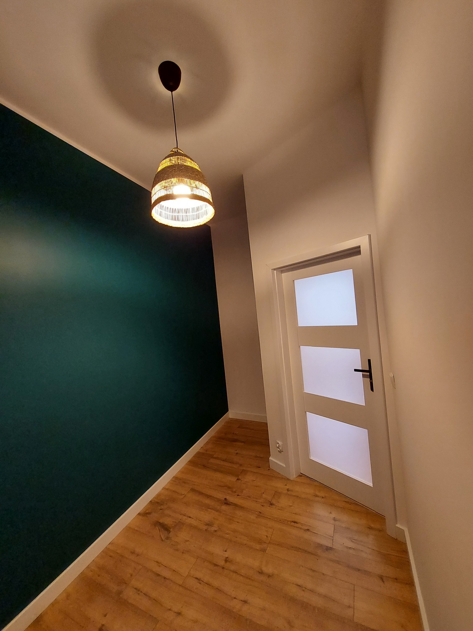 Wnętrze pokoju po remoncie: ciemnozielona ściana, drewniana podłoga, białe drzwi z trzema szybami i pleciona lampa wisząca.