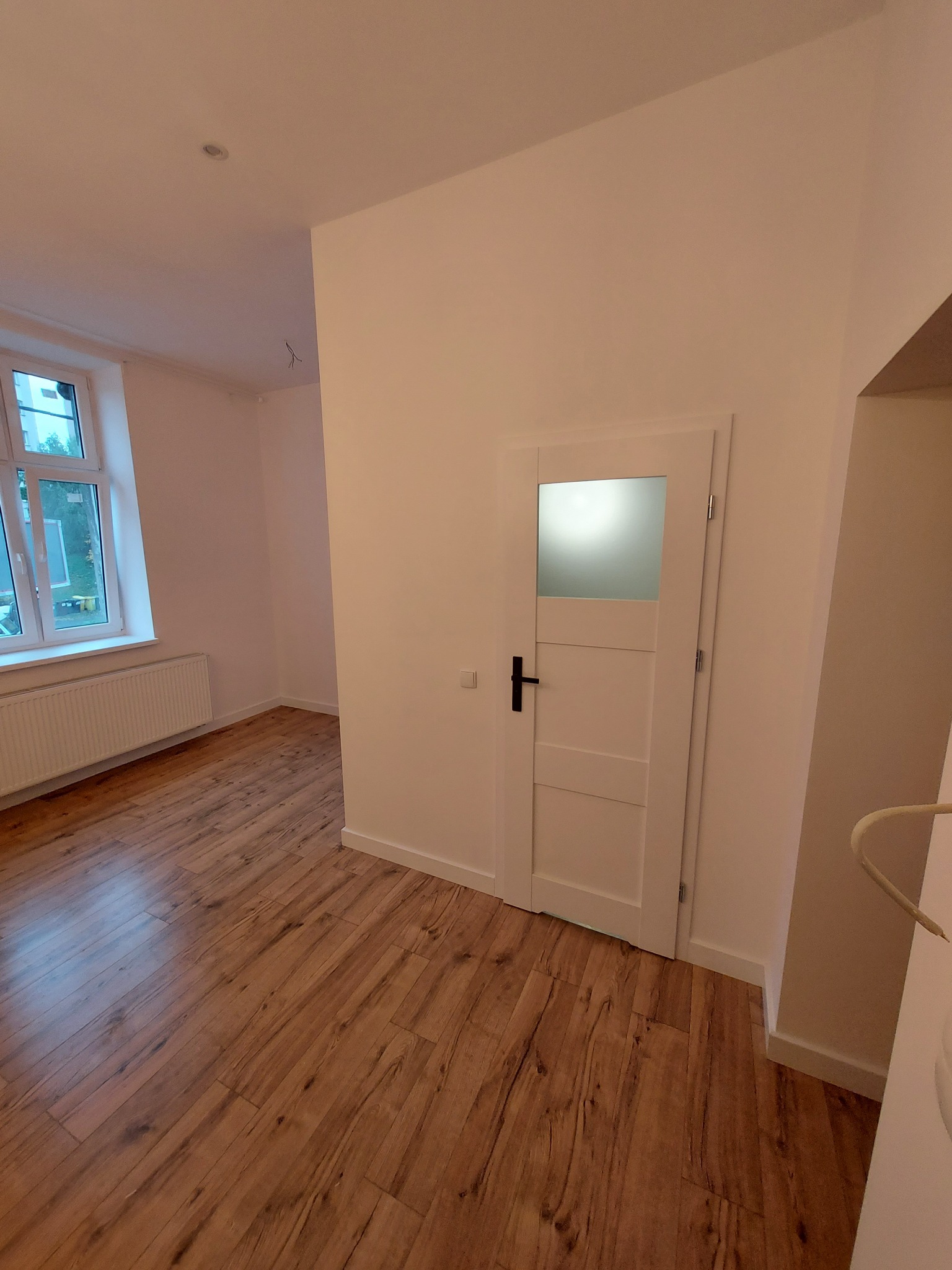 Pusty pokój po remoncie z drewnianą podłogą, białymi ścianami i drzwiami z przeszkleniem w górnej części, widok na okno z grzejnikiem.