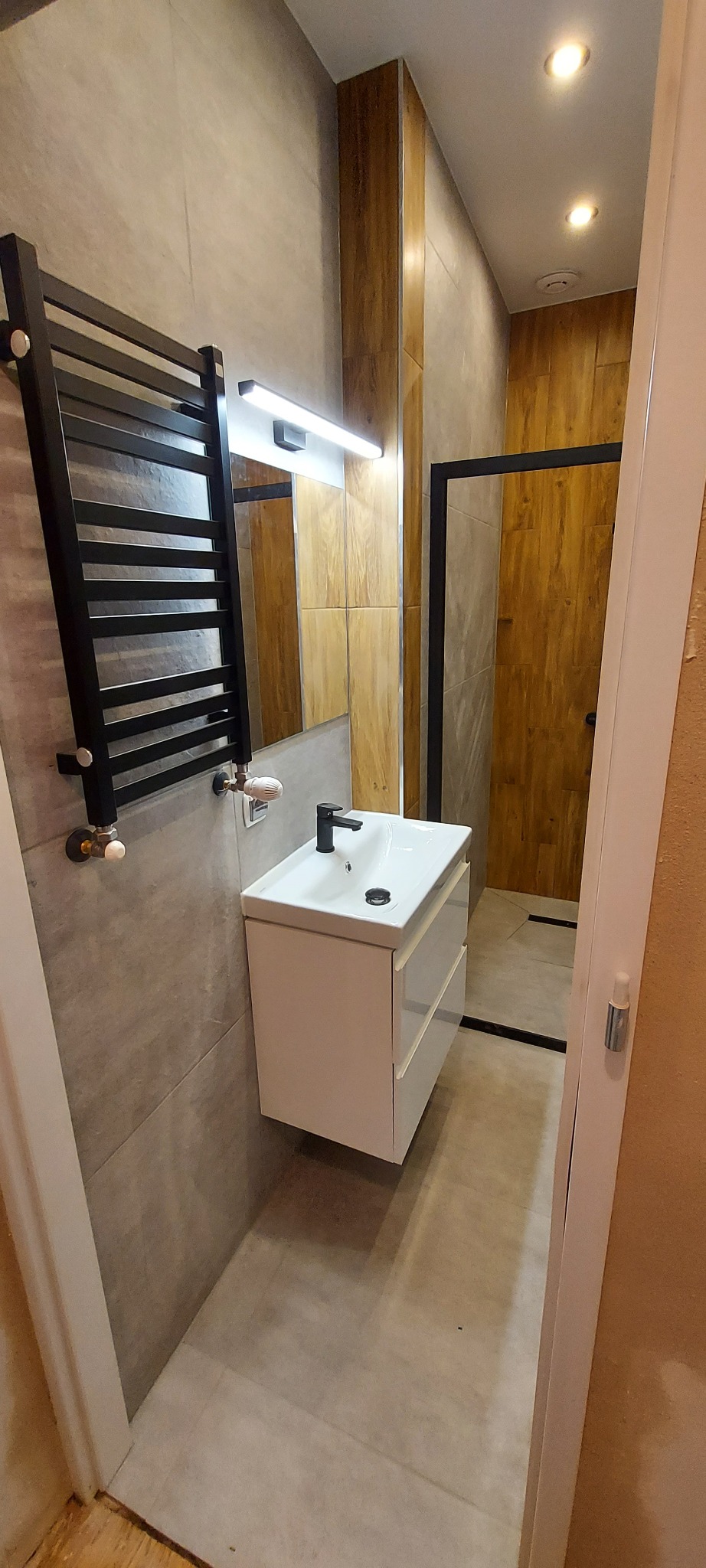 Nowoczesna łazienka z szarą płytką imitującą beton, drewnopodobnymi panelami na ścianie w strefie prysznica, białą szafką pod umywalką z czarną armaturą i czarnym grzejnikiem drabinkowym.