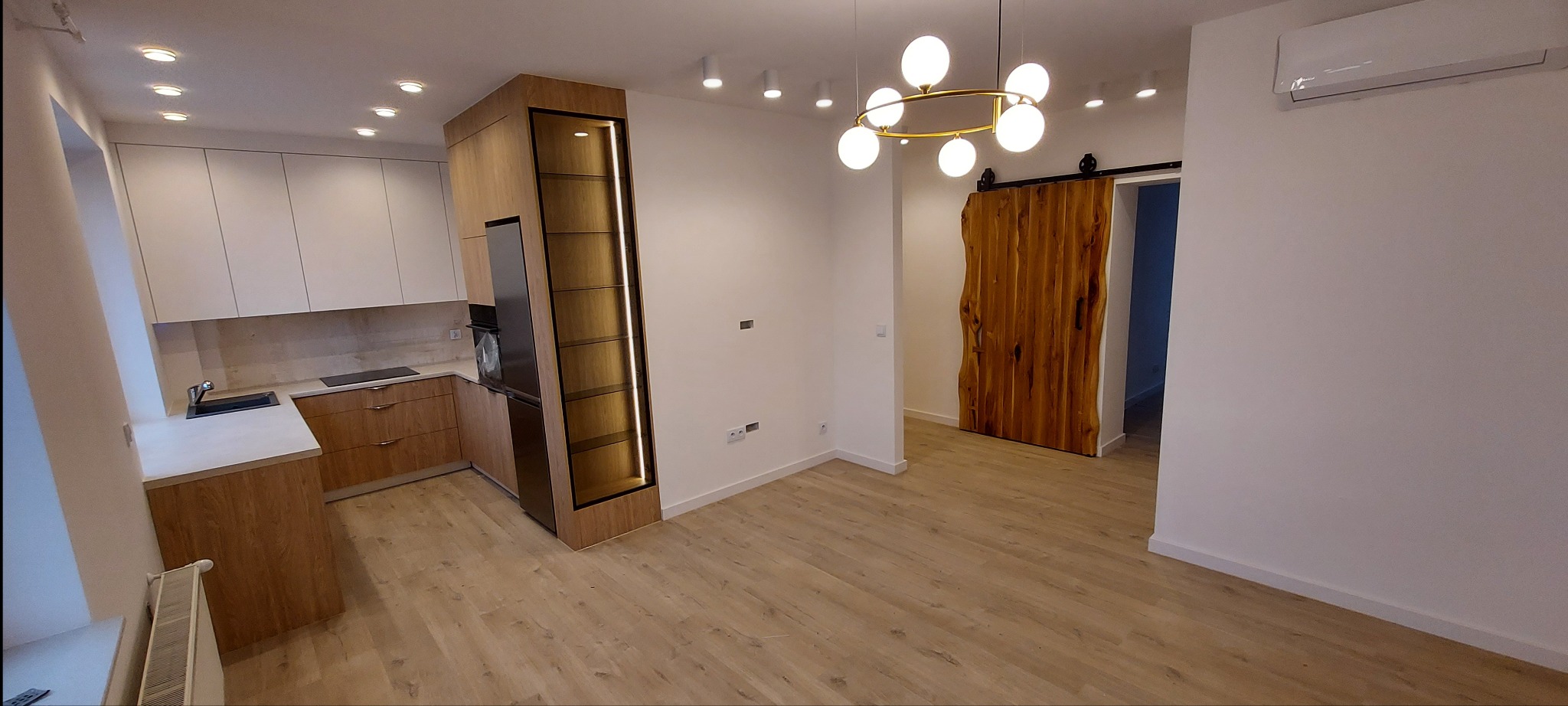 Nowoczesne wnętrze: kuchnia z drewnianymi szafkami i zabudowanym regałem z oświetleniem LED, widoczna drewniana przesuwna brama do innego pomieszczenia, oświetlenie sufitowe punktowe i okrągły...