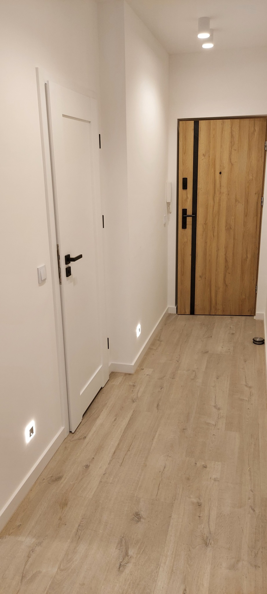 Nowoczesny przedpokój w Bytomiu z drewnopodobną podłogą, białymi ścianami i dwiema parami drzwi: wewnętrznymi białymi z czarną klamką i zewnętrznymi drewnianymi z czarnymi wstawkami.