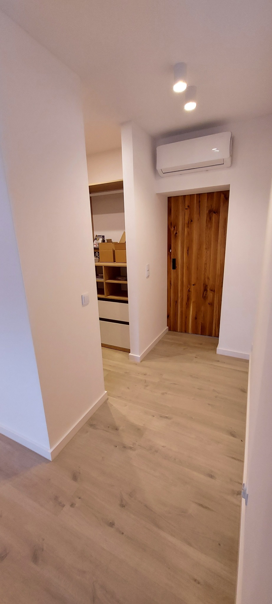 Wnętrze korytarza z drewnianą podłogą imitującą panele, białe ściany, drewniane drzwi przesuwne i klimatyzacja podsufitowa oraz widok na garderobę z półkami.