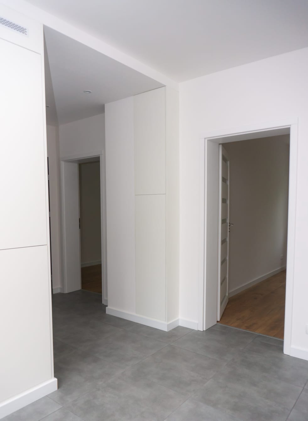 Jasne wnętrze mieszkania z szarymi płytkami podłogowymi, białymi ścianami i zabudową meblową, widok na otwarte drzwi do sąsiednich pomieszczeń z drewnianą podłogą.