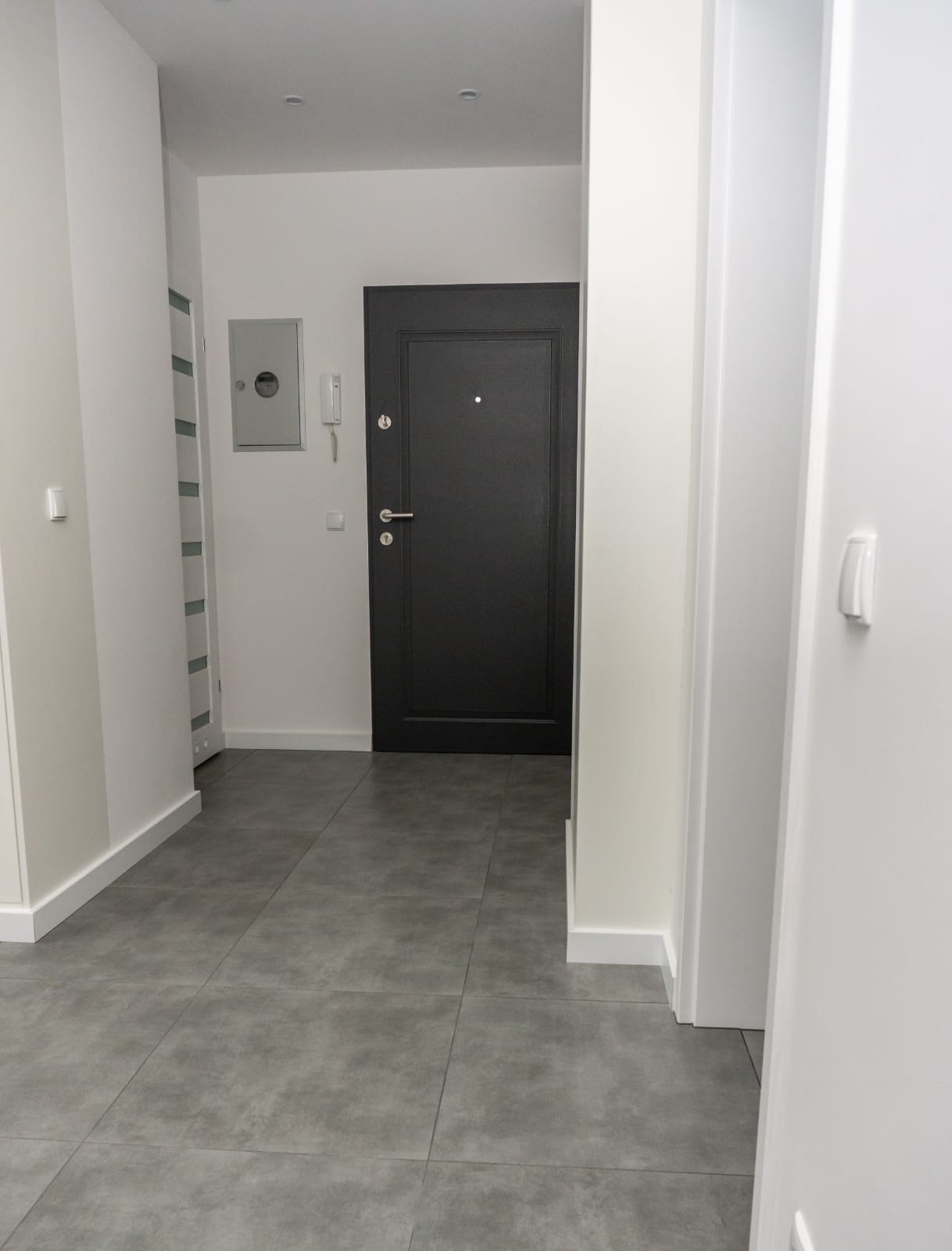 Nowoczesny przedpokój z szarą, kwadratową podłogą z płytek, białymi ścianami i ciemnymi drzwiami wejściowymi z metalowymi okuciami.