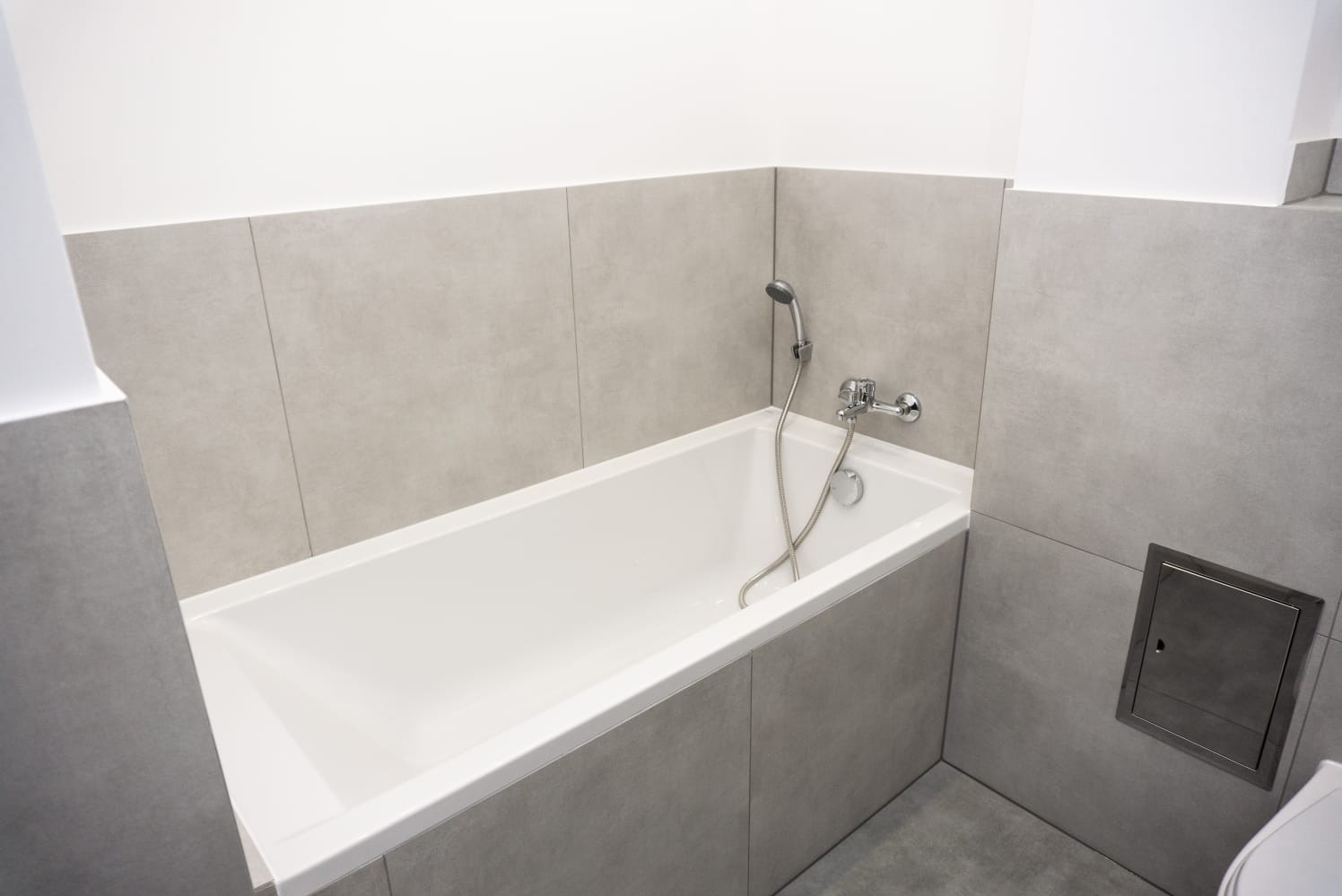 Nowoczesna, biała wanna obudowana szarymi płytkami ceramicznymi, z chromowaną baterią i słuchawką prysznicową, widoczny fragment klapy rewizyjnej w ścianie.