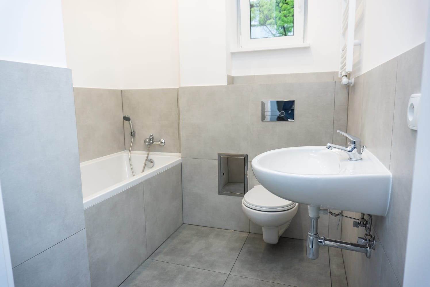 Jasna łazienka z wanną, umywalką narożną i toaletą w zabudowie, wykończona szarymi płytkami imitującymi beton, z oknem w tle.