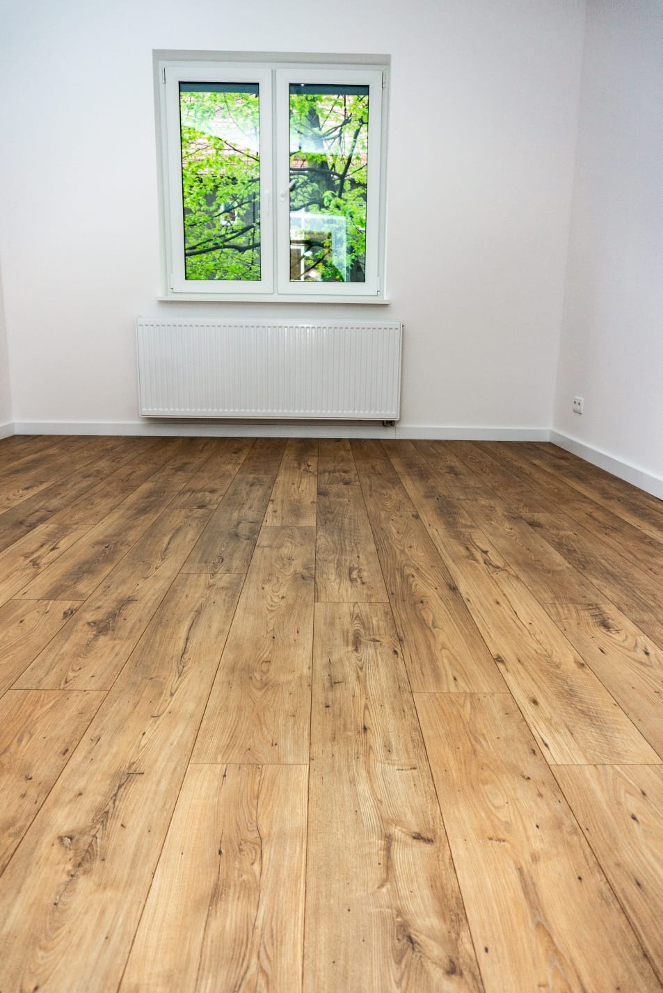 Jasna, drewnopodobna podłoga laminowana ułożona w pustym pokoju z białymi ścianami, grzejnikiem i oknem z widokiem na zieleń drzew.
