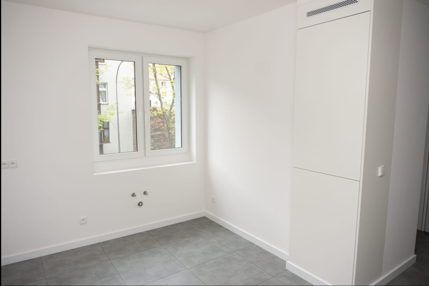 Puste, białe wnętrze pokoju z szarymi płytkami podłogowymi, białym oknem z widokiem na drzewo i budynek, oraz wbudowaną białą szafą z wentylacją u góry. Na ścianie widoczne gniazdka elektryczne...