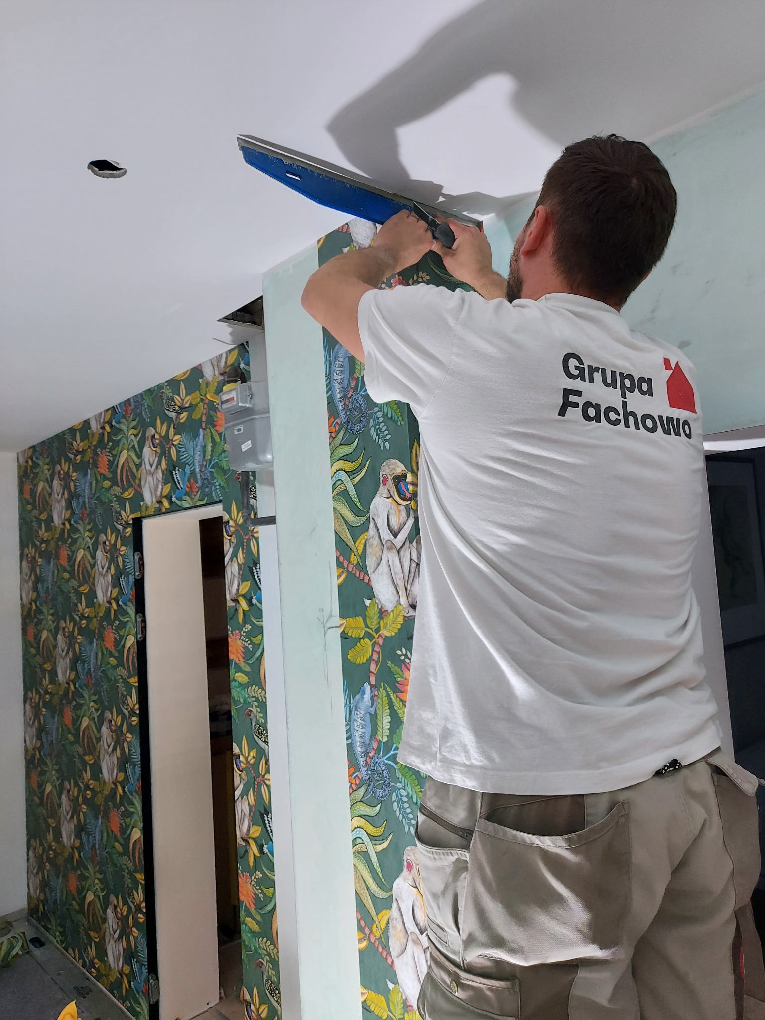 Pracownik w koszulce firmowej wygładza gładź na suficie w pomieszczeniu z tapetą w motywy dżungli.