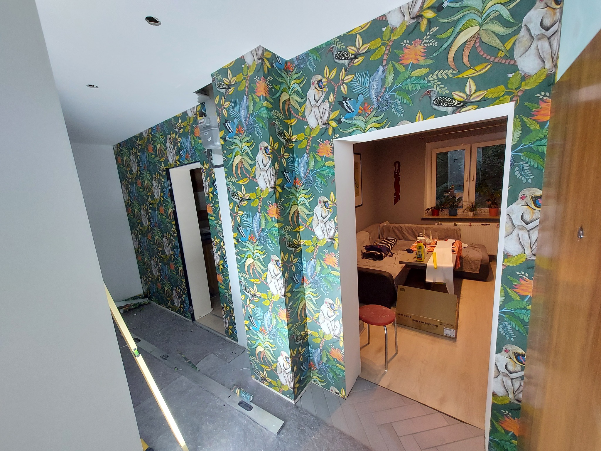 Wnętrze mieszkania w trakcie tapetowania ścian wzorzystą tapetą z motywem dżungli i małp, widoczne narzędzia i poziomica, fragment umeblowania w tle.