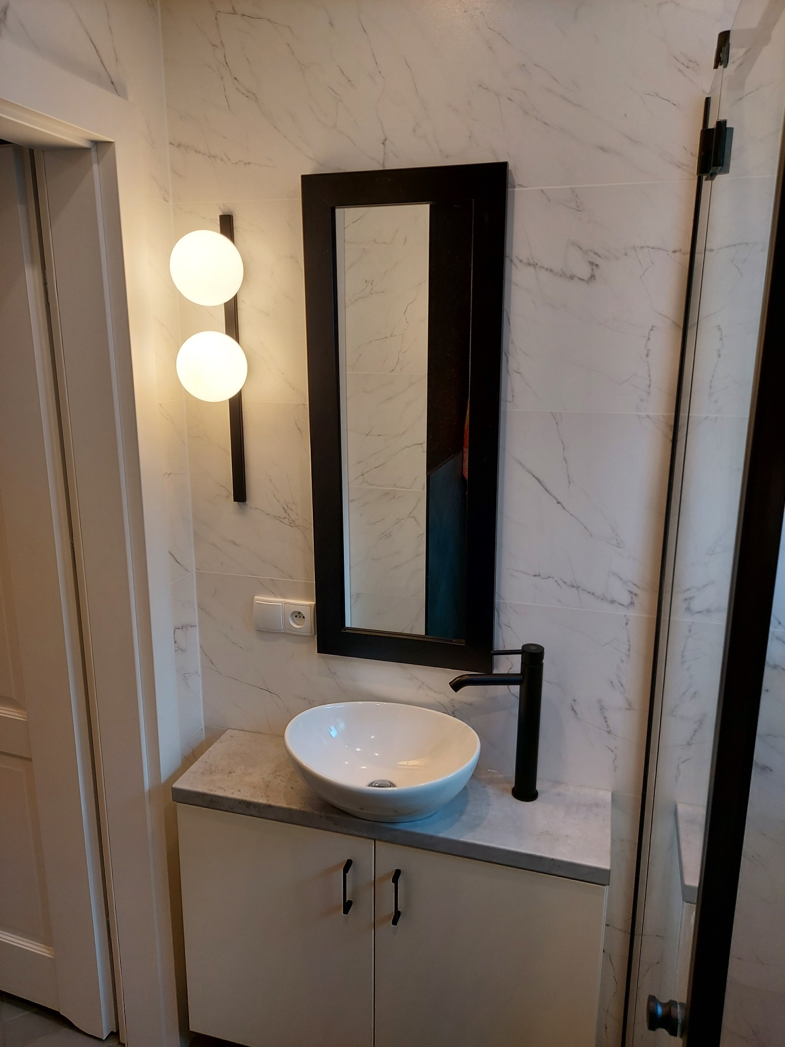 Nowoczesna łazienka z umywalką nablatową, czarną armaturą i podświetlanym lustrem na tle marmurowej ściany.