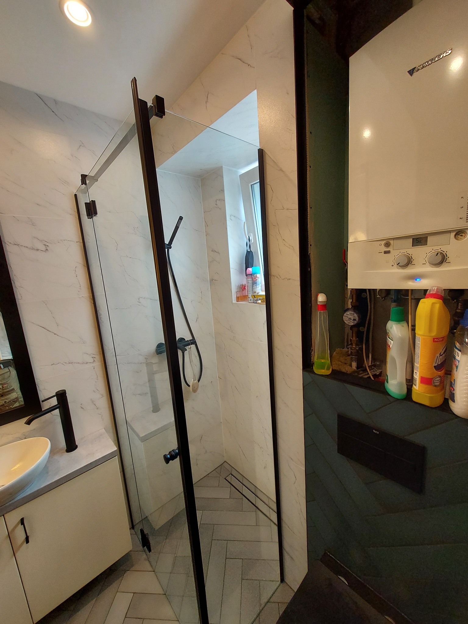 Nowoczesna łazienka z kabiną prysznicową typu walk-in z czarnymi profilami, umywalką nablatową na szafce w kremowym kolorze, marmurowymi płytkami na ścianach i podłodze ułożonej w jodełkę; obok...